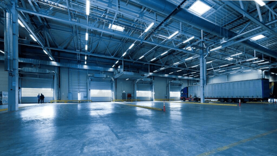 Inside a contract logistics warehouse space. There are two people talking near a closed roller door and there is a truck and trailer parked on the other side.