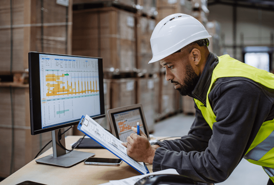 Person in warehouse looking at clipboard with data on the computer screen next to them.