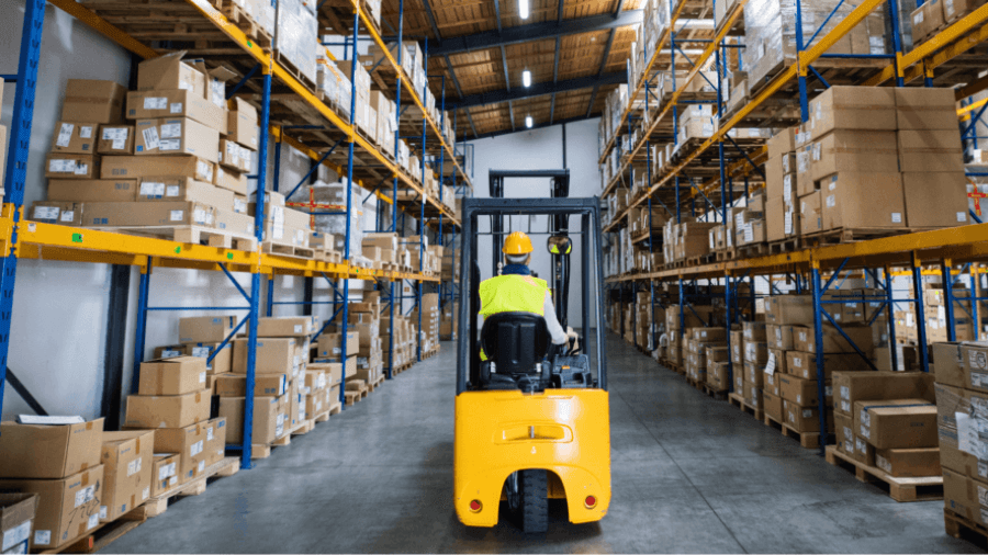 Person driving a forklift in warehouse, through aisles of pallet racking.