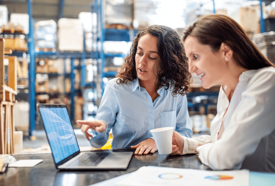 Two women looking at a laptop discussing warehouse rates. They are sitting at a desk within a warehouse.